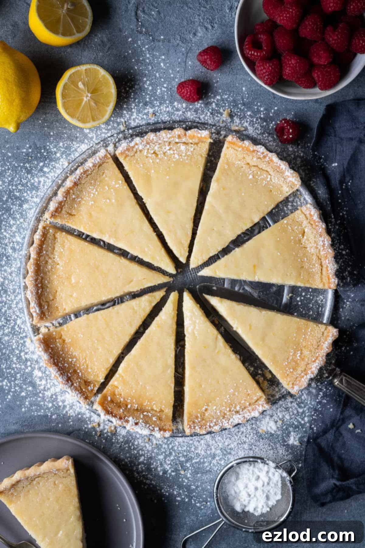 Sliced vegan lemon tart on a grey background with fresh lemons and vibrant raspberries.