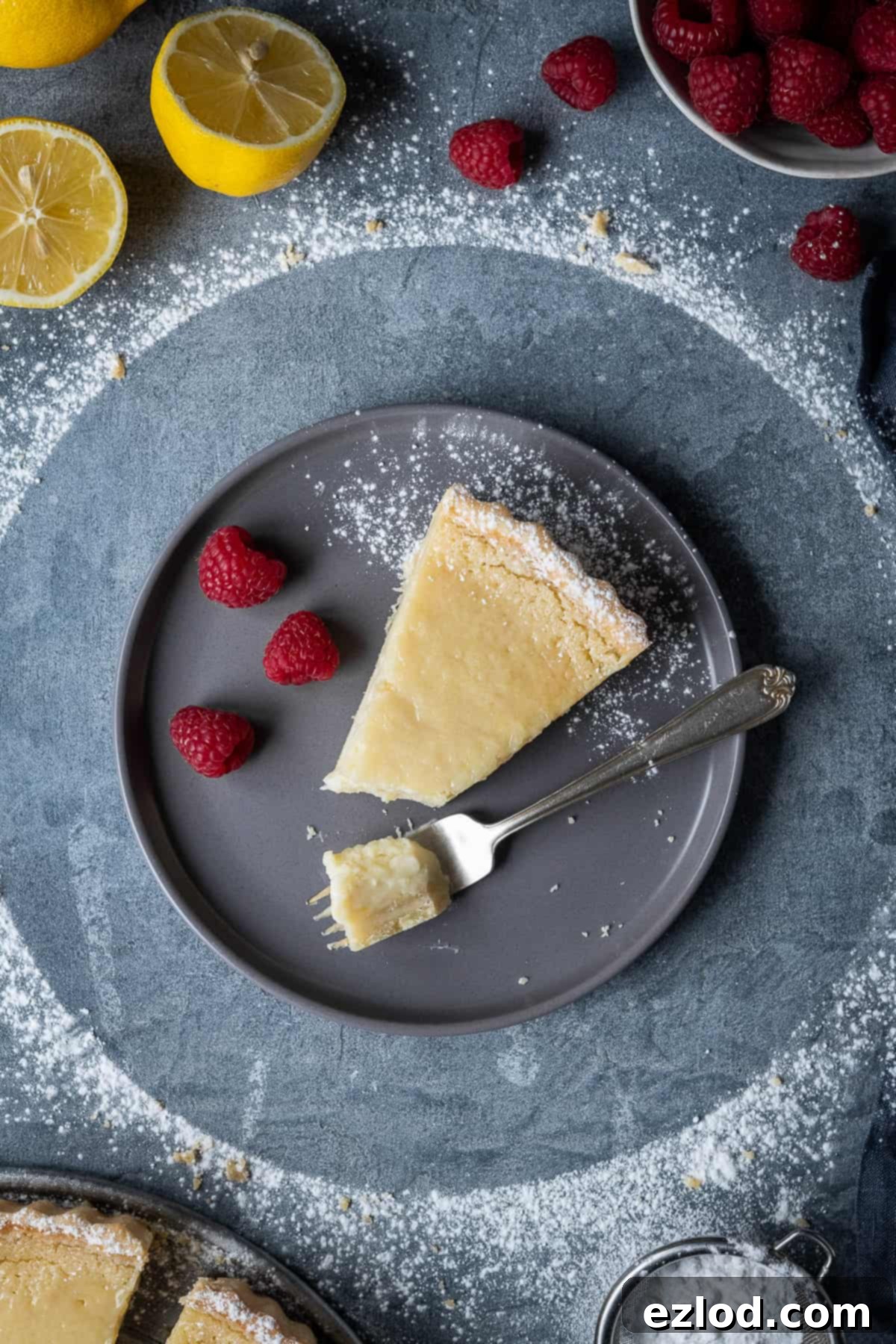 A slice of vegan lemon tart with fresh raspberries and a fork on a grey plate, ready to be eaten.