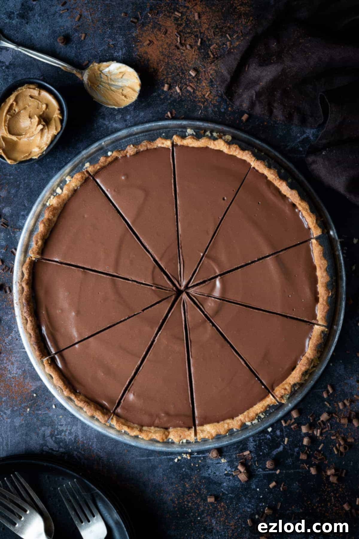 A beautifully sliced vegan peanut butter chocolate tart on a dark background, garnished with delicate chocolate curls. Plates, forks, and a bowl of creamy peanut butter accompany it, creating an inviting scene.
