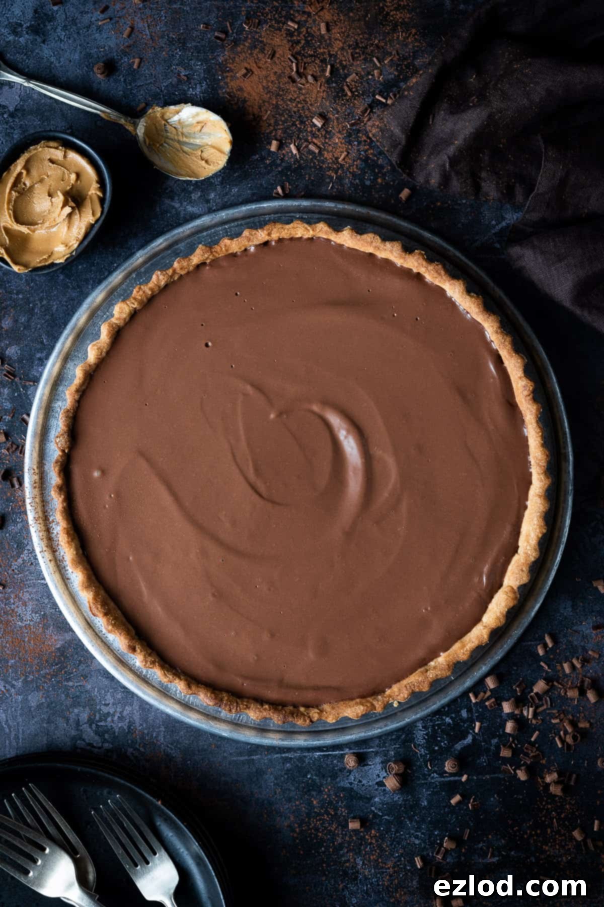 A complete vegan peanut butter chocolate tart on a rustic metal tray, surrounded by chocolate shavings, a bowl of peanut butter, and elegant cutlery on a dark, textured background.