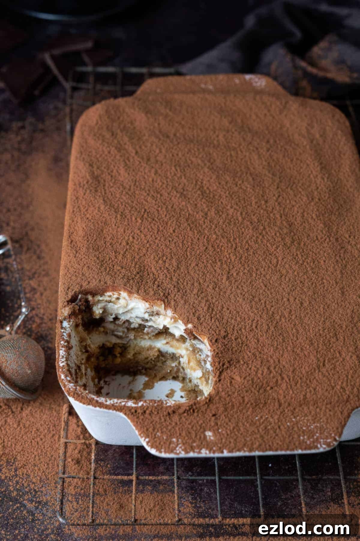 A white dish of vegan tiramisu with a spoonful removed on a wire rack on a dark background dusted with cocoa powder. This rich, creamy, and irresistible dessert is a perfect vegan baking project.
