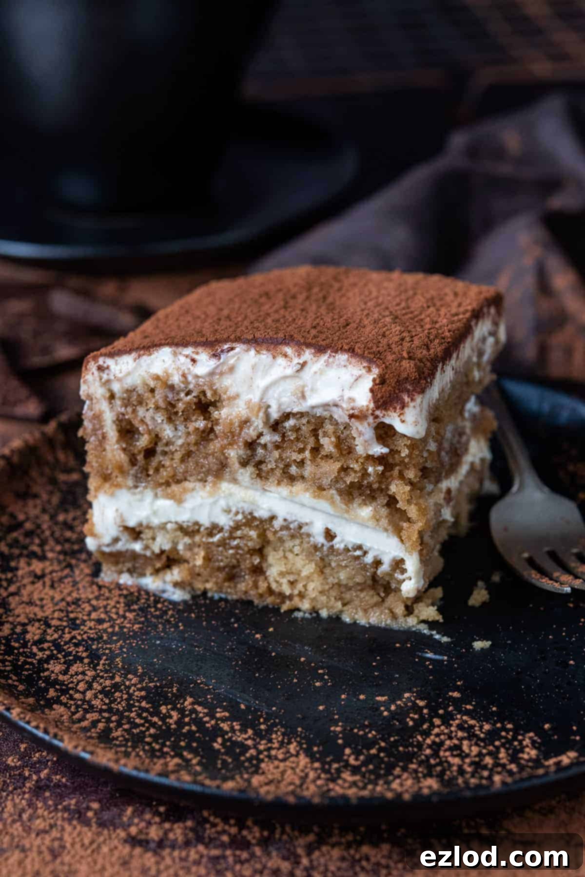 A slice of vegan tiramisu on a black plate with a fork, showing its rich layers of cake and cream.