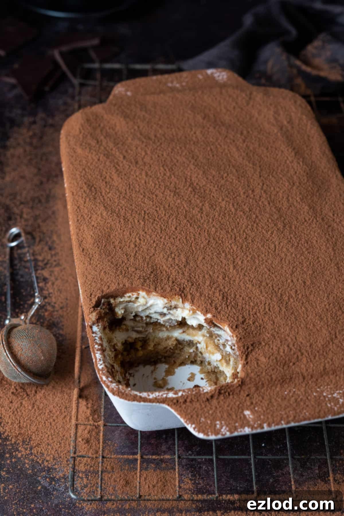 A white dish of vegan tiramisu on a dark background, garnished with cocoa powder, dark chocolate shavings, and a brown cloth.