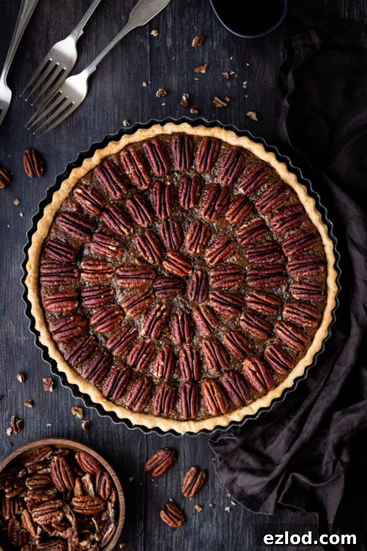 Vegan pecan pie on a wooden backdrop with a bowl of pecans, forks and a brown cloth.