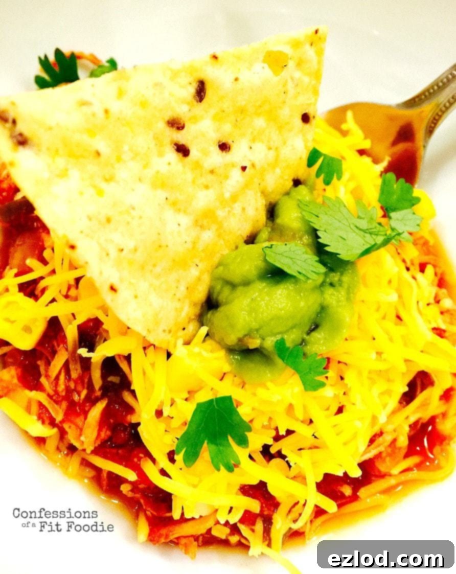 Crock Pot Chili Chicken topped with cheddar cheese, fresh cilantro, sliced avocado, and a single tortilla chip dipped in the middle. A fork rests on the side of the bowl, showcasing a hearty, healthy meal.