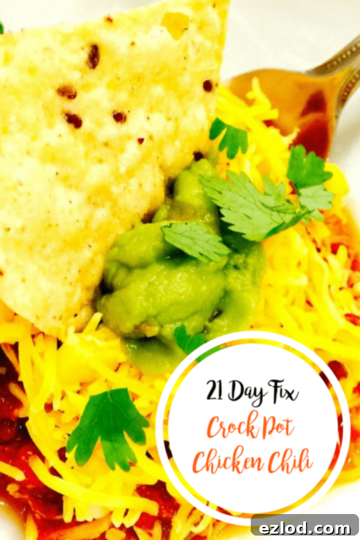 A vibrant bowl of 21 Day Fix Crock Pot Chicken Chili, generously topped with melted cheddar cheese, fresh cilantro, and creamy avocado slices. A single tortilla chip is perfectly dipped in the center, inviting you to dig in.