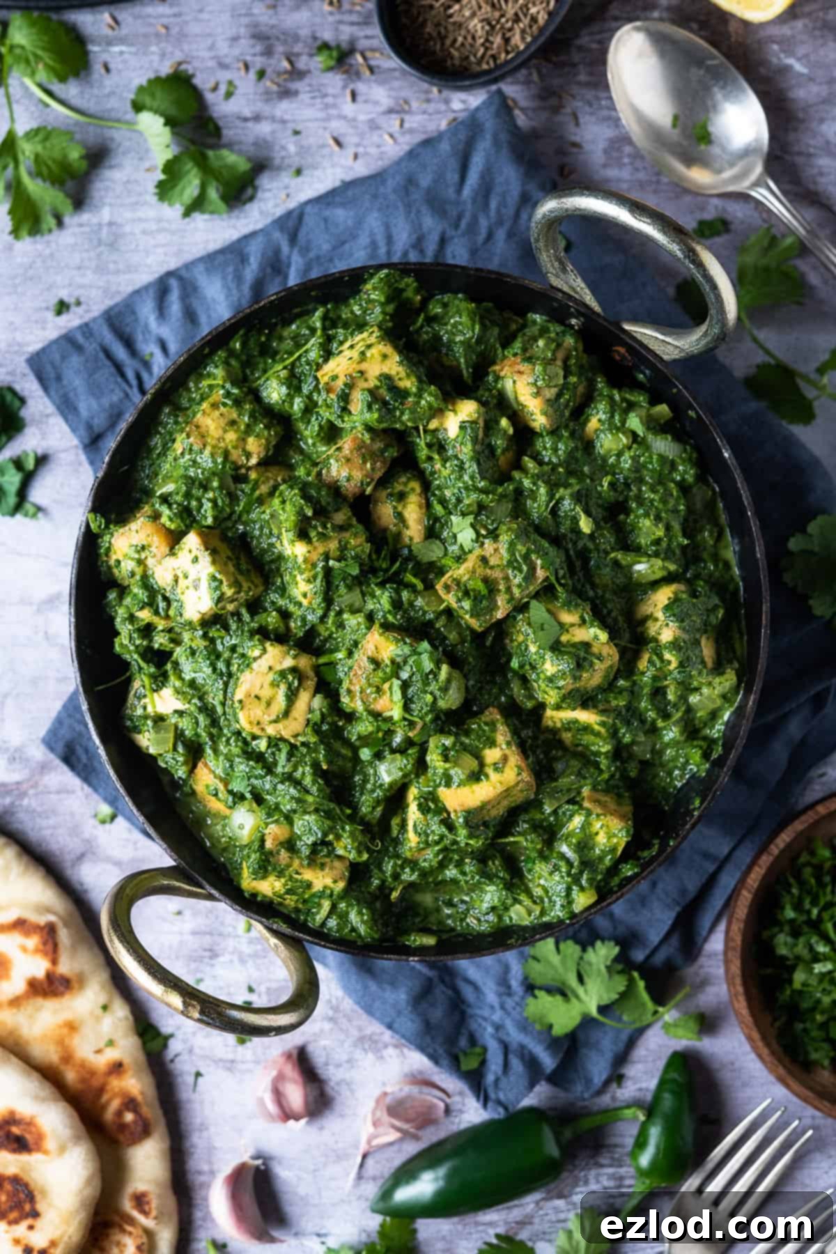 Vibrant Vegan Palak Paneer in a traditional kadhai pan, surrounded by fluffy naan bread, fresh coriander, green chillies, and aromatic garlic cloves, set against a stylish grey surface. A perfect representation of a delicious plant-based Indian meal.