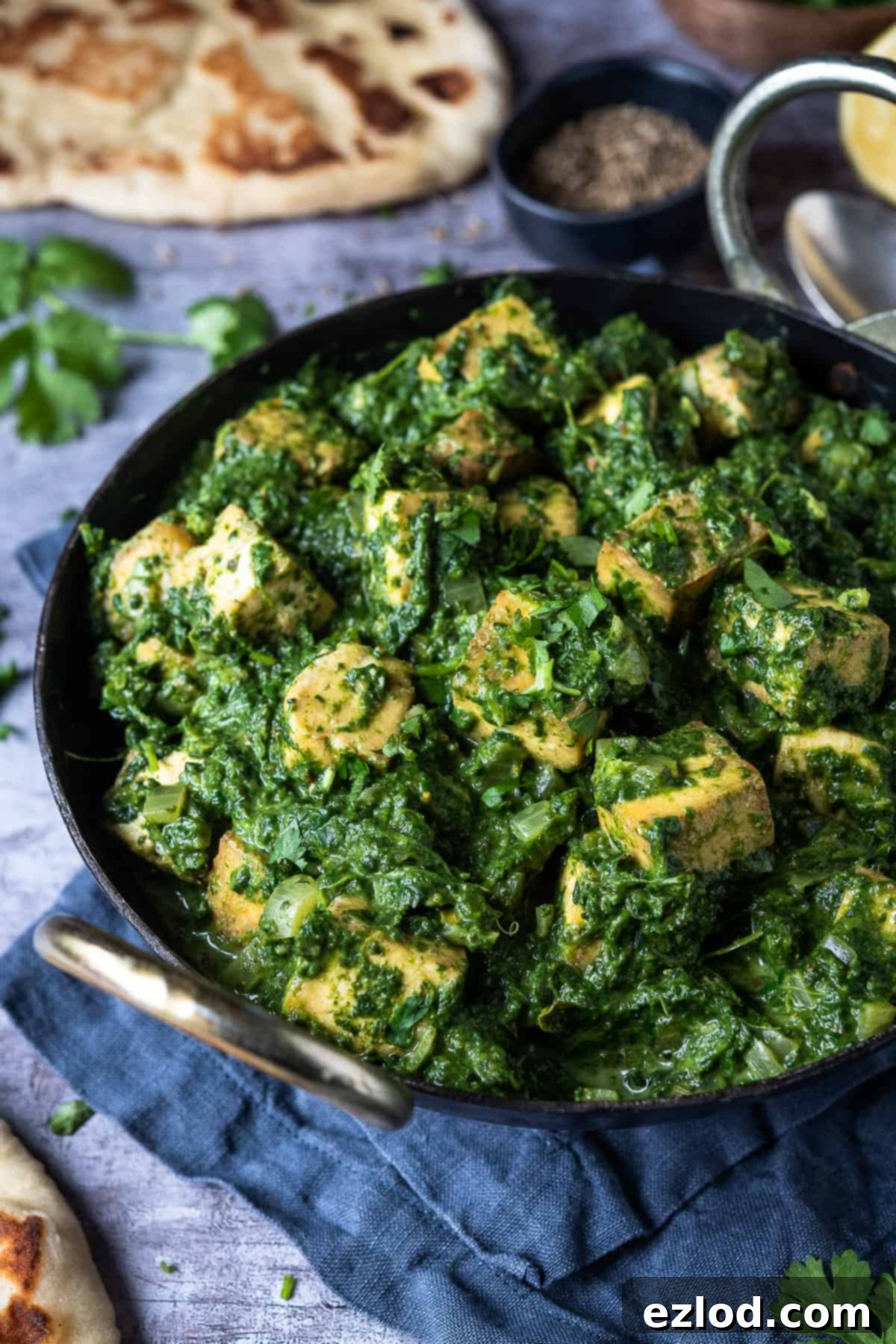 A delicious bowl of Vegan Palak Paneer, featuring creamy spinach gravy and golden tofu cubes, served in a black kadhai pan with soft naan bread visible in the background, all on a sleek grey surface.