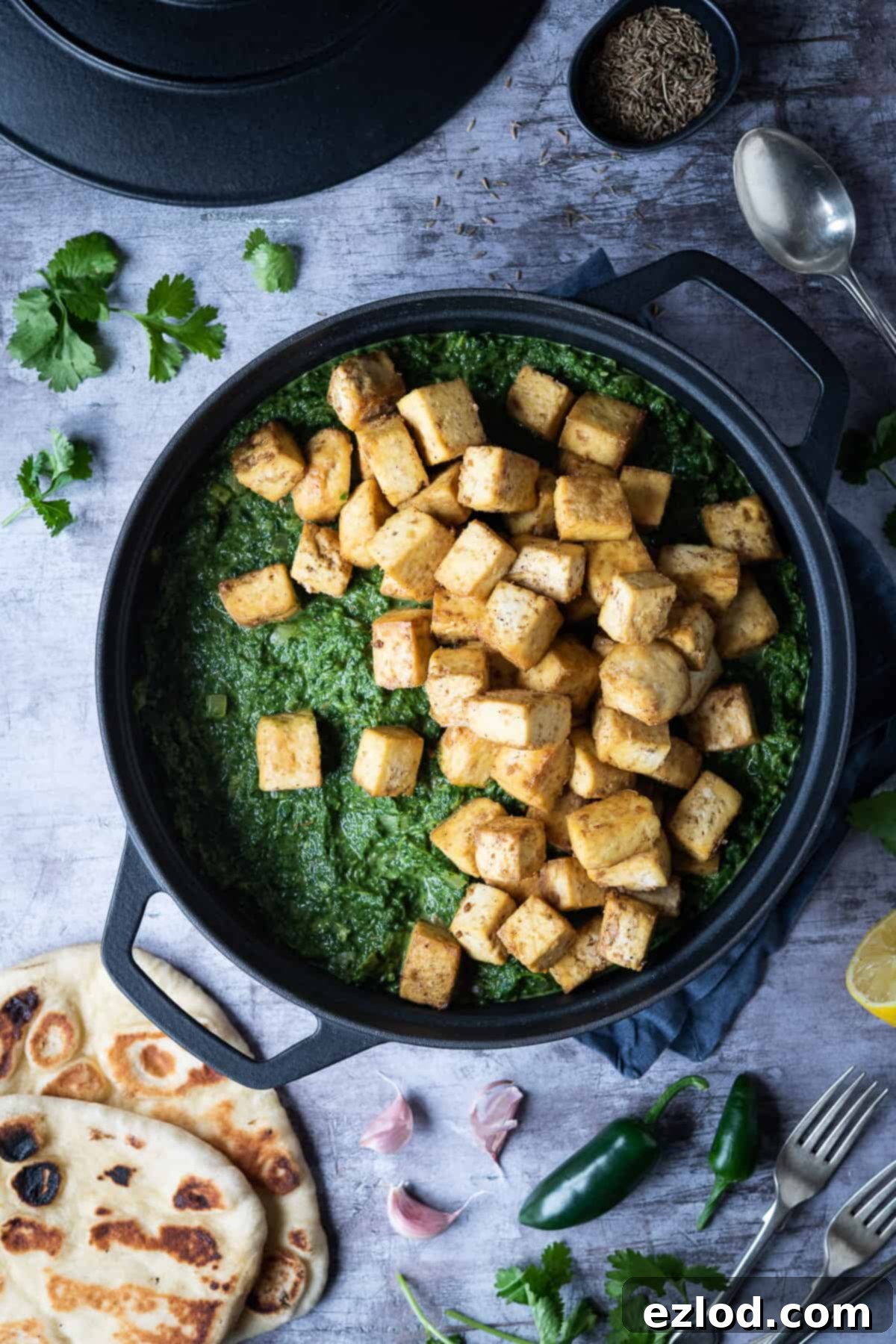 Perfectly baked tofu 'paneer' cubes resting on a bed of luscious, vibrant green spinach sauce in a black saucepan, ready to be gently folded in and served as a delicious Vegan Palak Paneer.