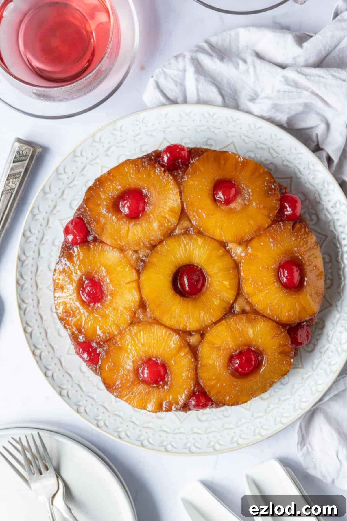 A beautifully presented vegan pineapple upside-down cake on a white plate, set against a chic marble background. Accompanying it is a delicate cup of fruit tea, additional plates, and a soft grey cloth, creating a cozy and inviting scene.
