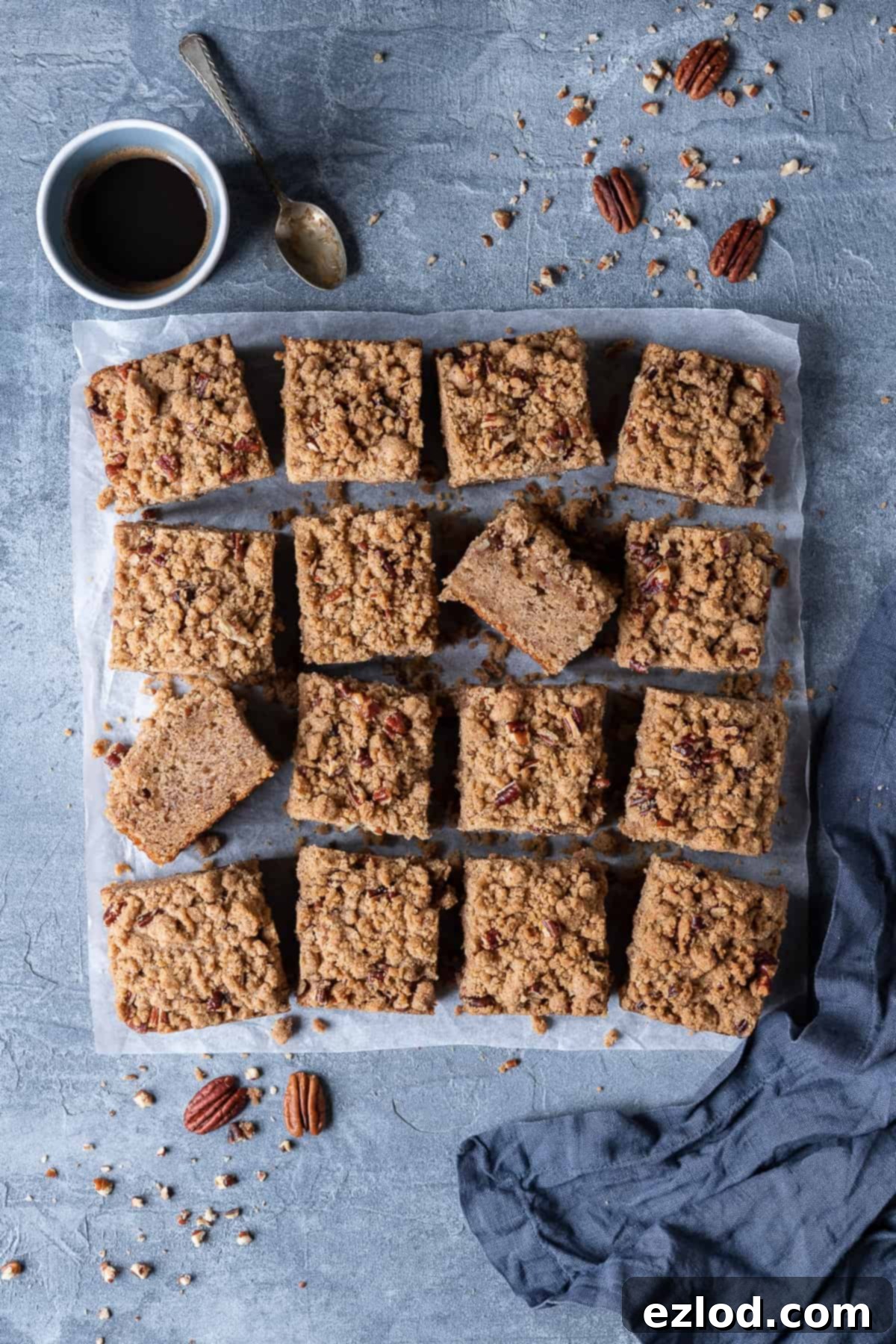 Vegan banana crumb cake sliced into squares on a grey background with a cup of espresso, a grey cloth and chopped pecans.