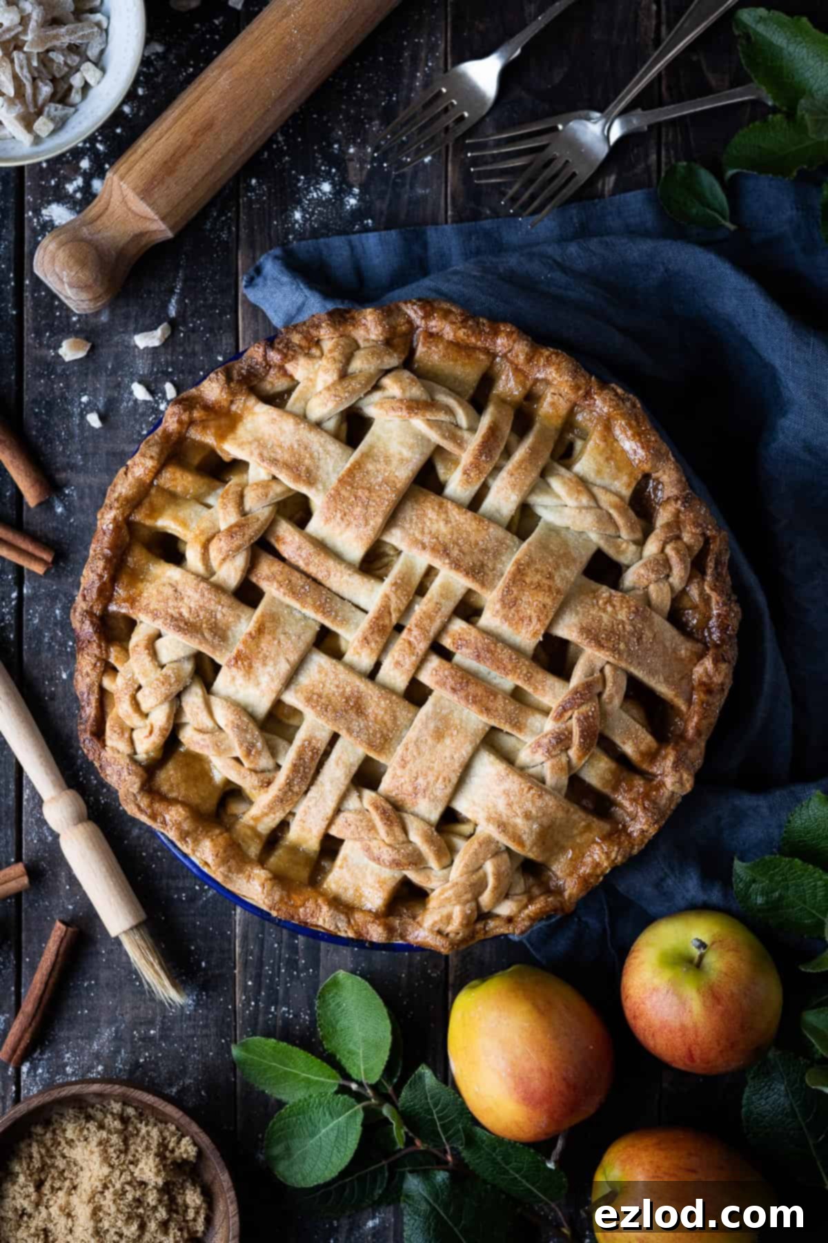 A beautifully baked vegan ginger apple pie on a dark wooden table, surrounded by fresh apples, a linen cloth, baking ingredients like flour and sugar, and kitchen equipment. The pie has a golden-brown lattice crust.