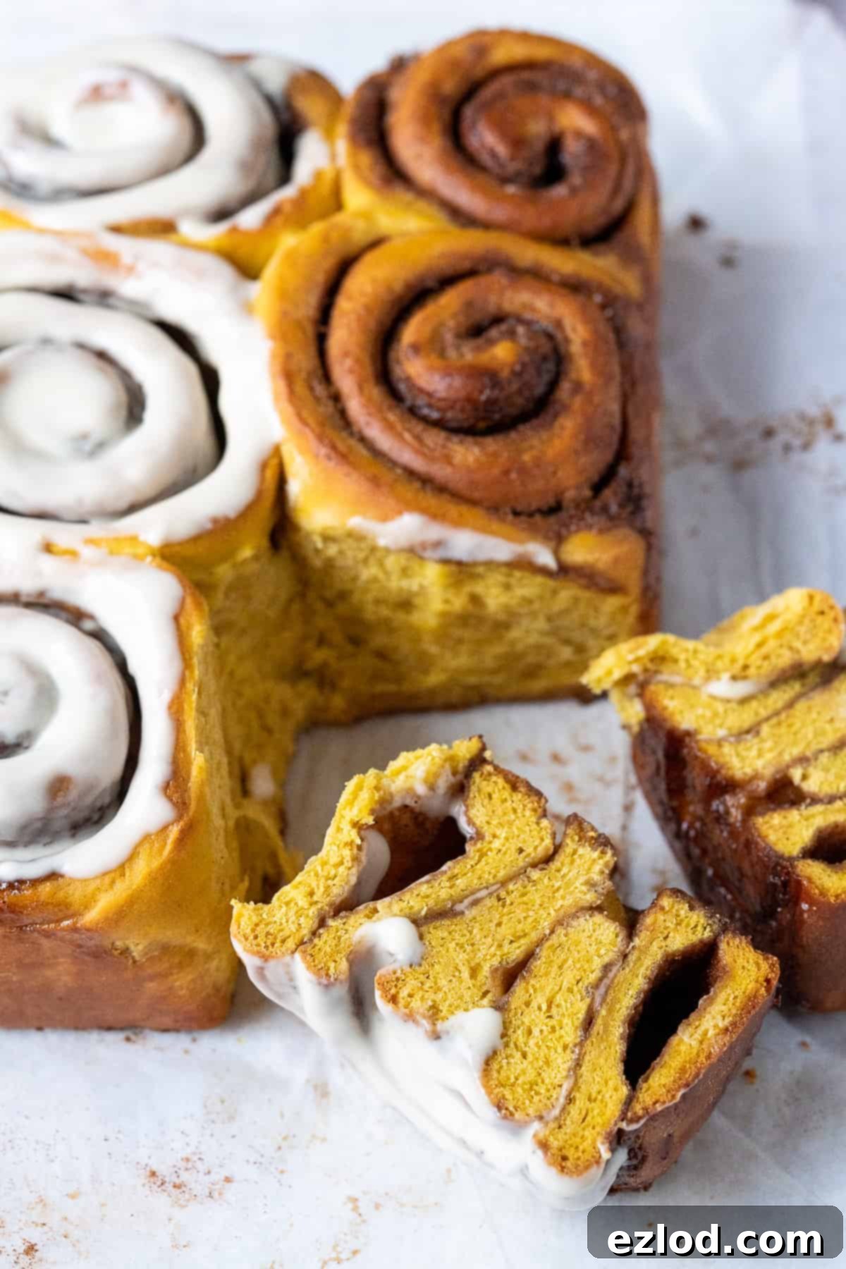 Delicious vegan pumpkin cinnamon rolls on a piece of white baking parchment, with one roll expertly sliced in half to reveal its fluffy texture and cinnamon swirl.