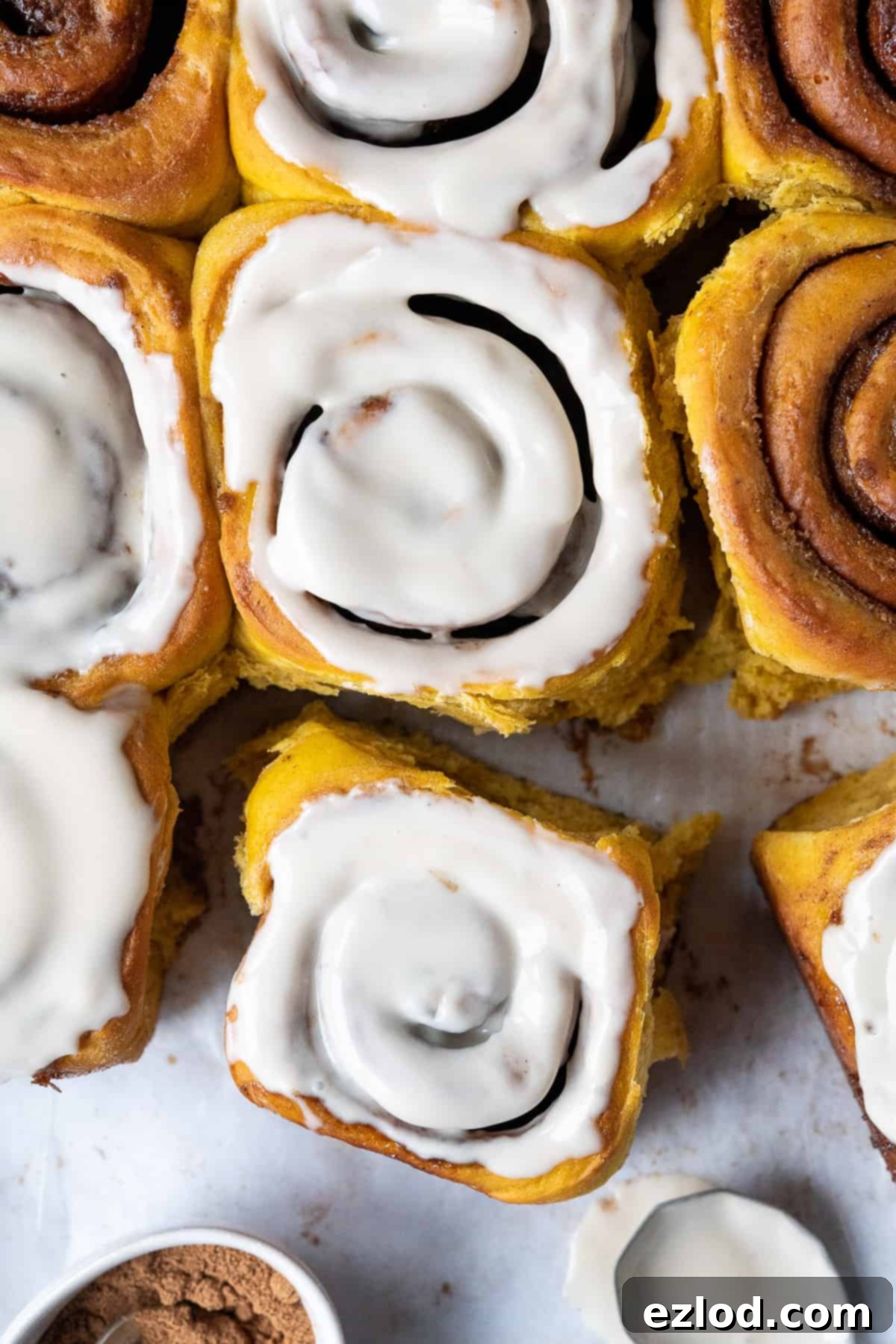 Vegan pumpkin cinnamon rolls, one drizzled with maple glaze, on white baking parchment with a small bowl of pumpkin spice and a spoon of glaze.