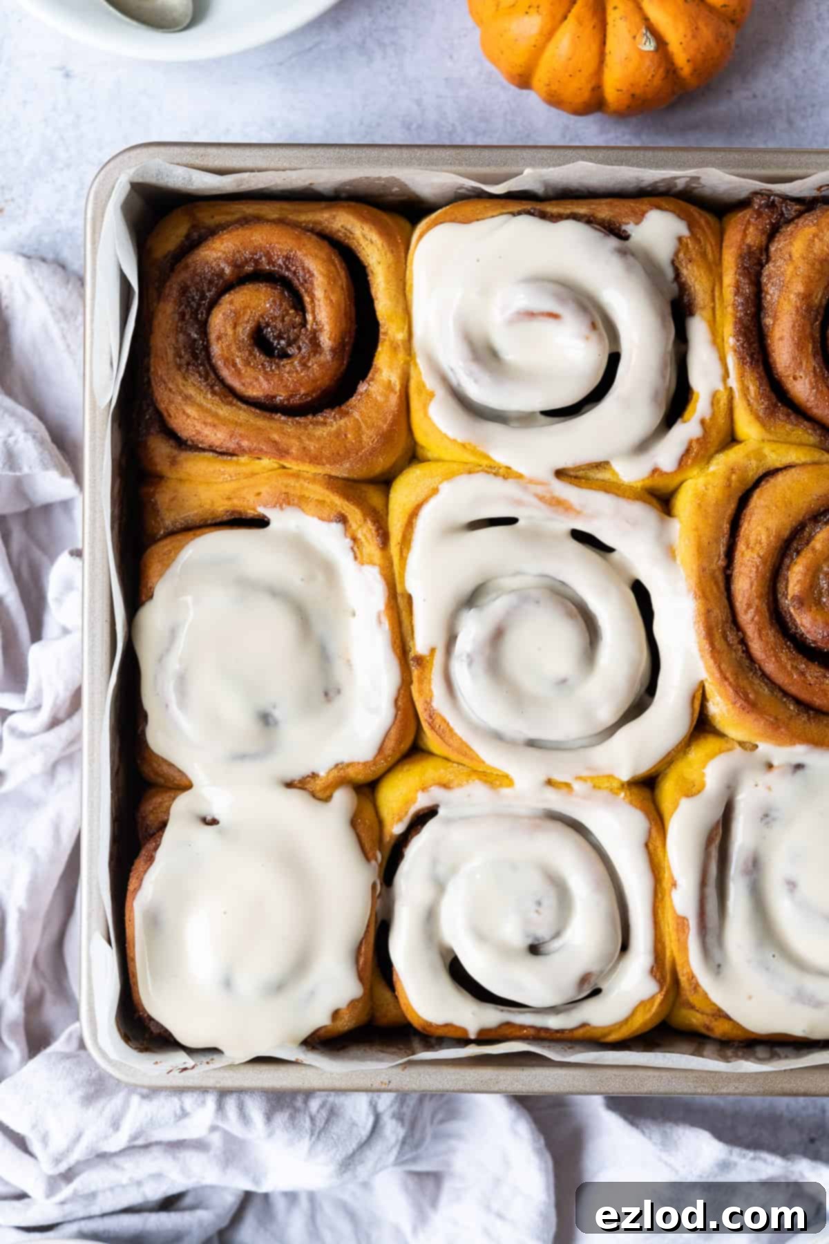 Beautifully glazed vegan pumpkin cinnamon rolls in a metal cake tin, with a soft grey cloth and a small decorative pumpkin, evoking a cozy autumn scene.