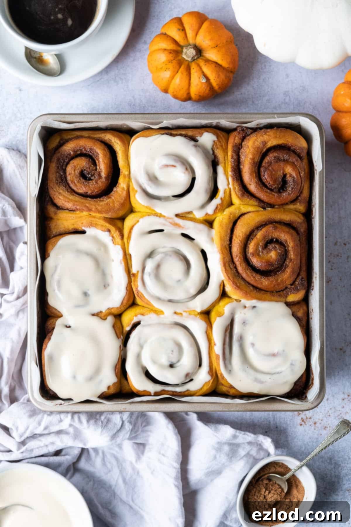 A baking tin filled with freshly baked vegan pumpkin cinnamon rolls, unglazed, resting on a rustic grey background. Mini decorative pumpkins and a warm cup of coffee are placed beside the tin, enhancing the cozy autumn theme.