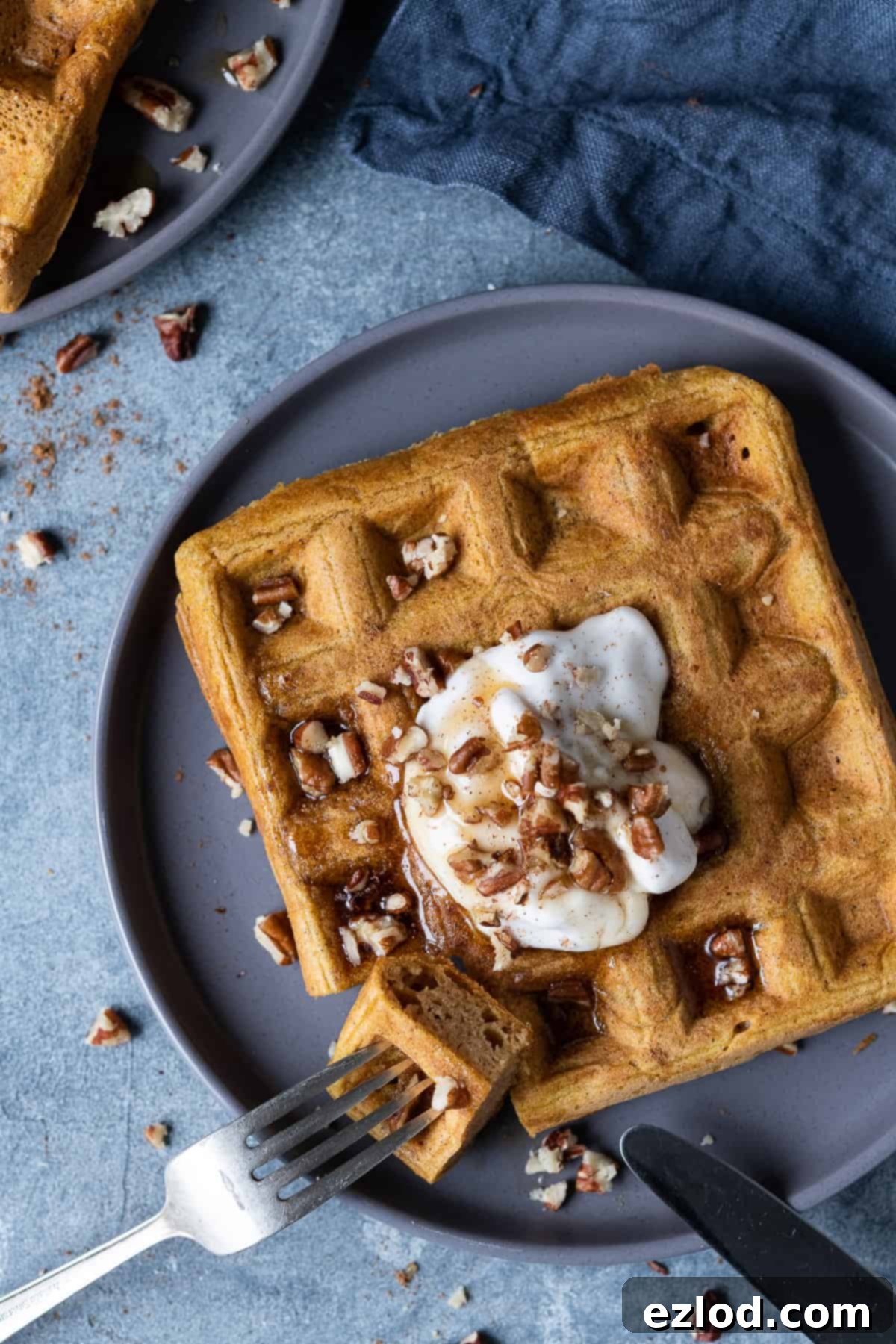 A vegan pumpkin waffle topped with yogurt, chopped pecans and maple syrup on a grey plate with a knife and fork.