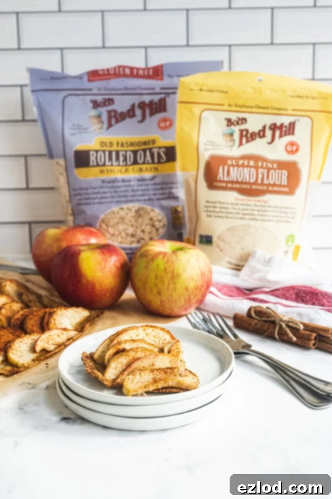 A beautifully presented slice of apple tart on a stack of plates in the foreground, with fresh apples, cinnamon sticks, and Bob's Red Mill oats and flour artfully arranged in the background, showcasing the key ingredients.