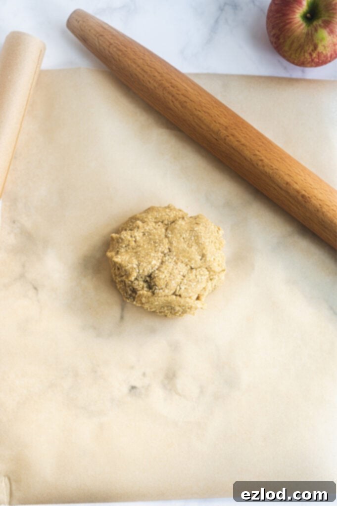 An overhead photo showcasing brown parchment paper with a neatly formed ball of Gluten-Free dough, accompanied by a classic wooden rolling pin, perfectly set for the next step of preparing the tart crust.