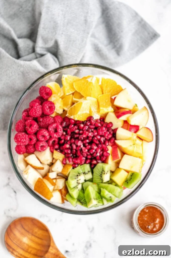 Fruits are diced and placed evenly in a large glass bowl on a white marble surface, near a wooden spoon and gray towel.