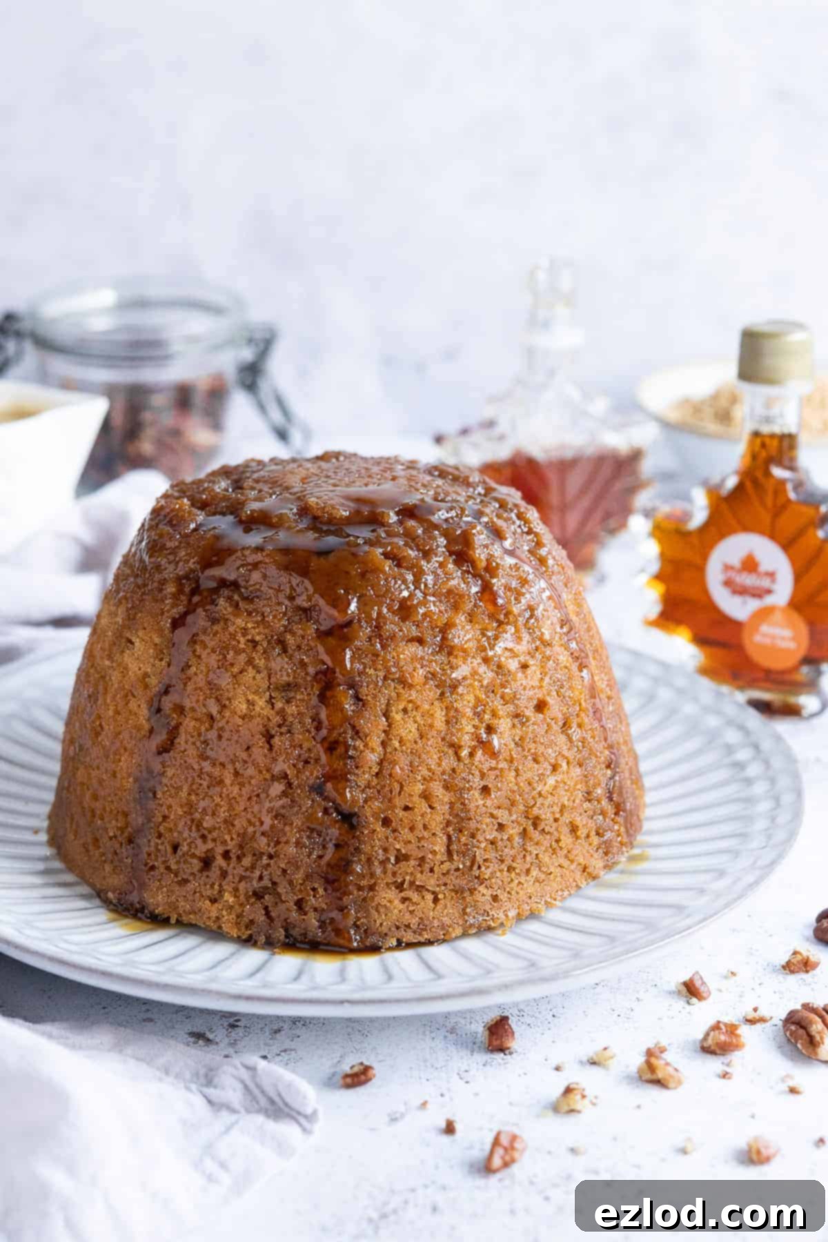 Vegan steamed maple syrup pudding on a white plate with bottles of maple syrup, a jug of custard, and a jar of pecans in the background, creating a rustic and inviting scene.