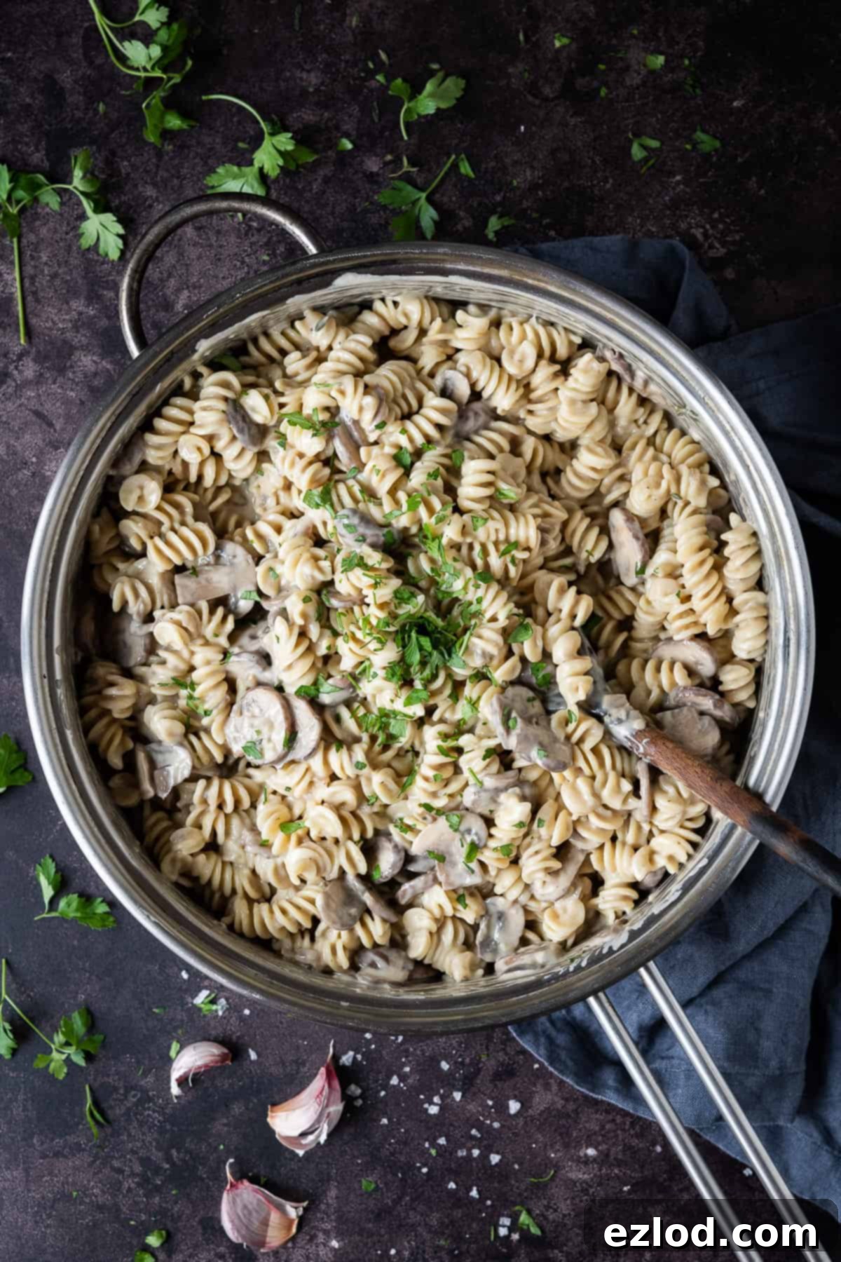 A metal pan of creamy vegan mushroom pasta on a dark surface with fresh parsley and garlic skins.