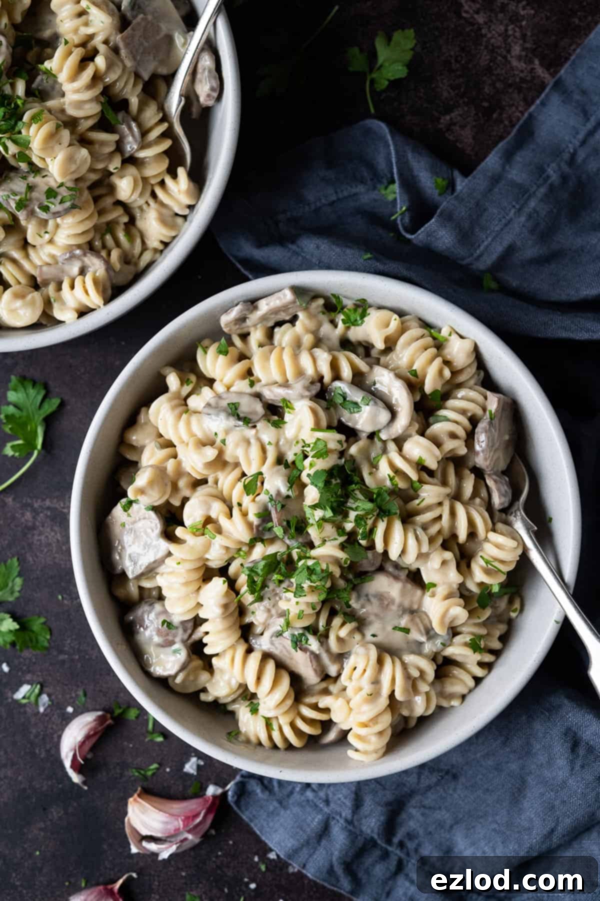 Two grey bowls of mushroom pasta on a dark background with a grey cloth, fresh parsley and garlic skins.