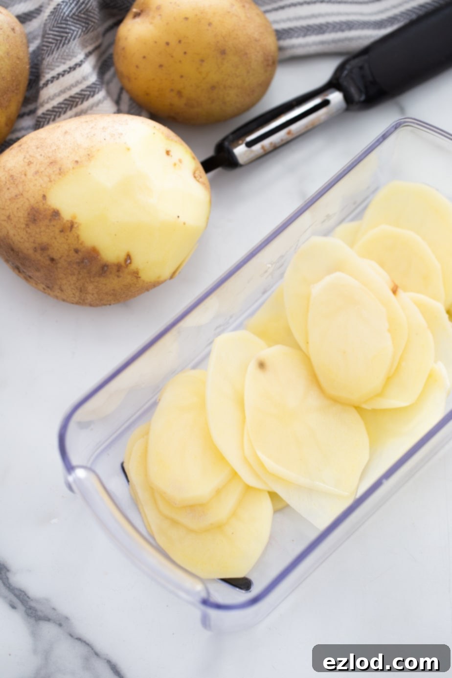 A hand peeling a potato next to a clear glass bowl filled with thinly sliced potatoes, ready for cooking.