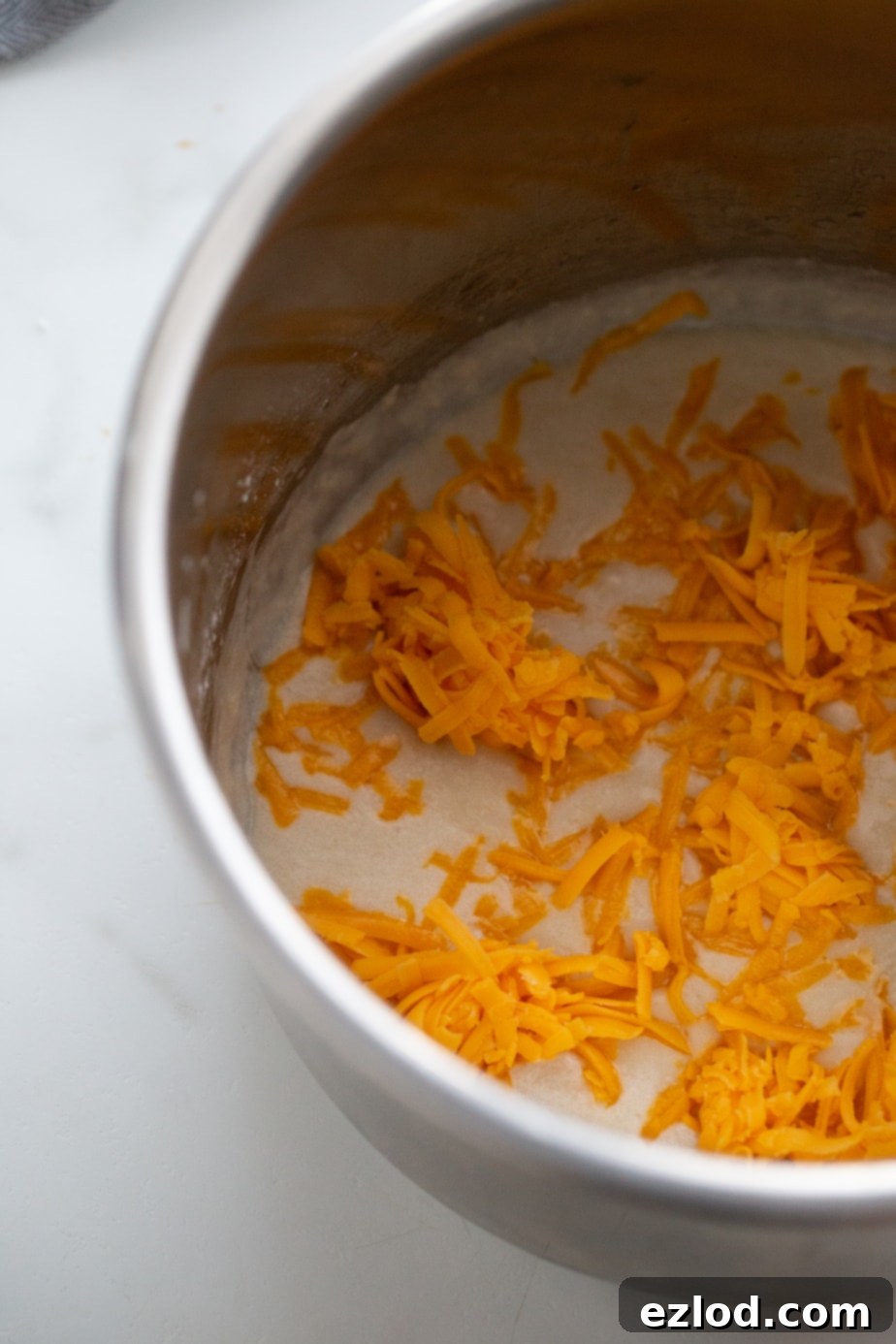 A pot on a stovetop with freshly shredded yellow cheddar cheese melting, ready to be incorporated into a creamy sauce.