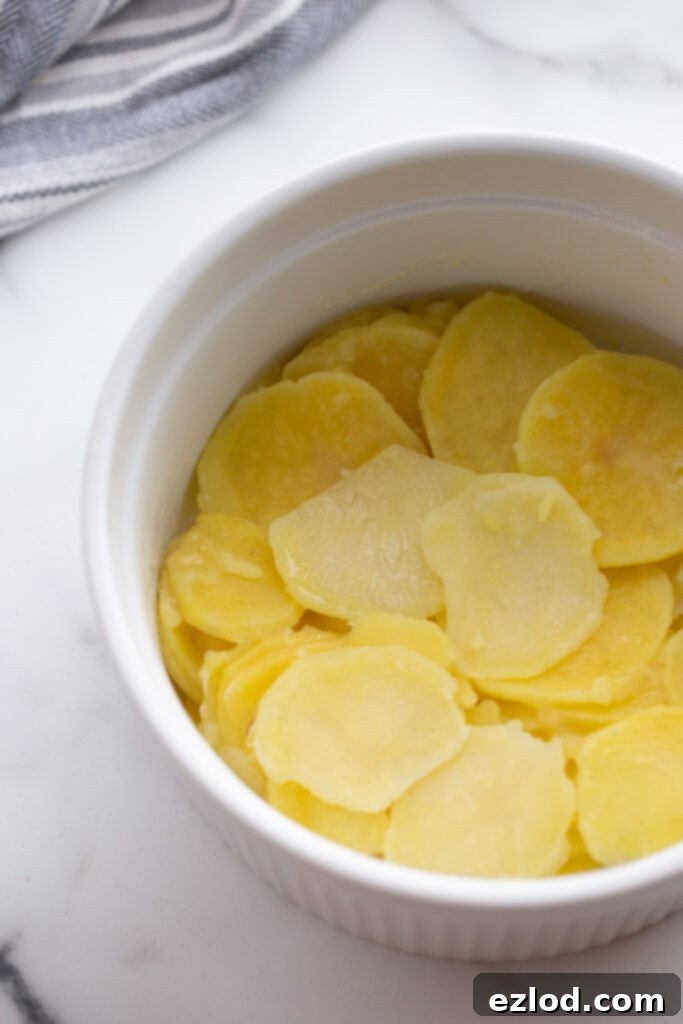 Thinly sliced yellow potatoes are neatly arranged in a white mixing bowl, lightly seasoned.