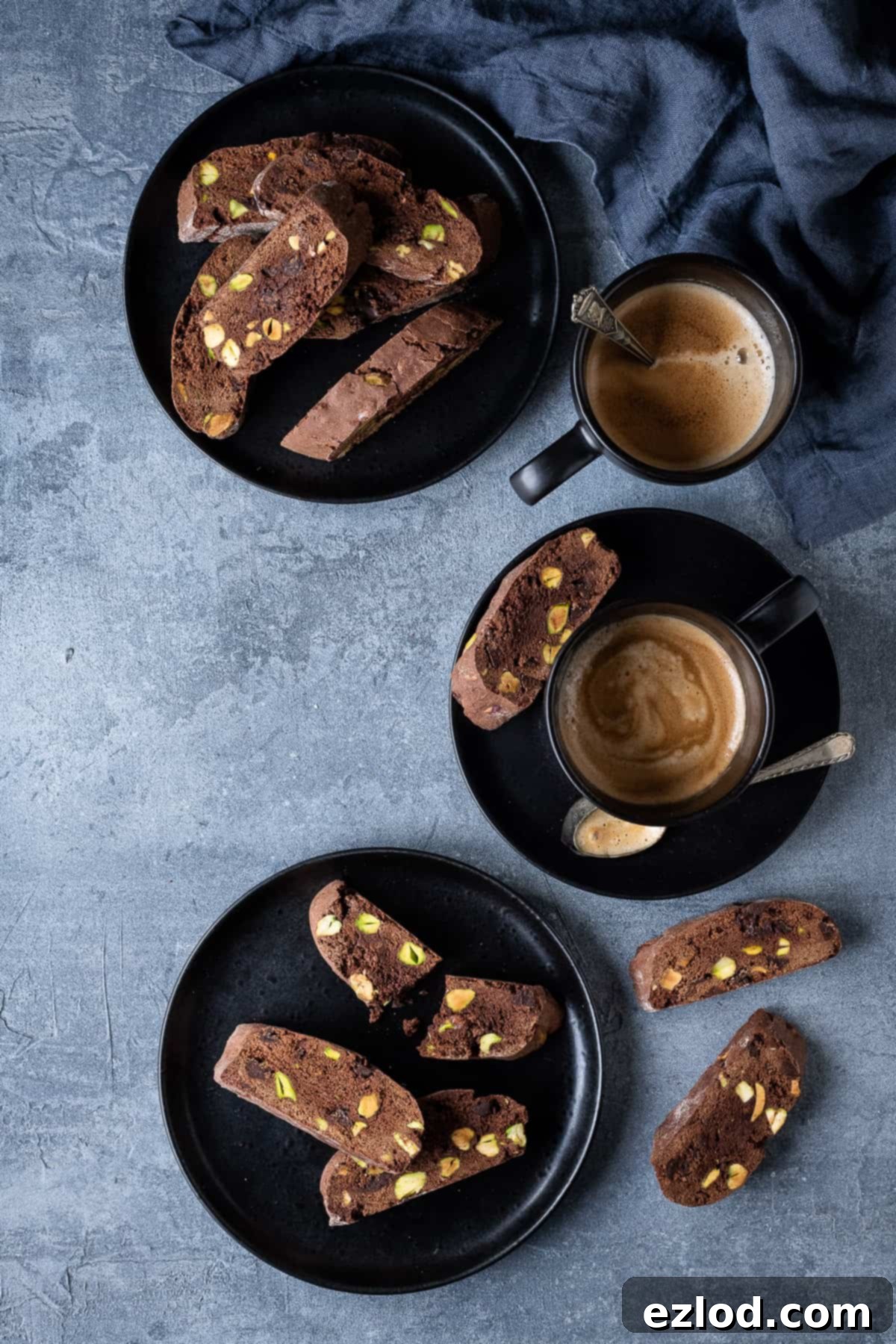 Chocolate pistachio biscotti neatly arranged on black plates, accompanied by black cups of espresso coffee and a soft grey cloth.