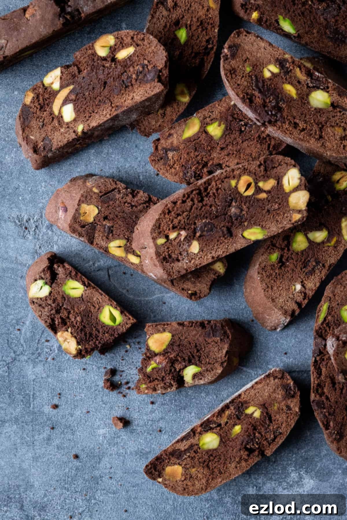 Several vegan chocolate pistachio biscotti on a grey background, with one broken in half to reveal its interior.