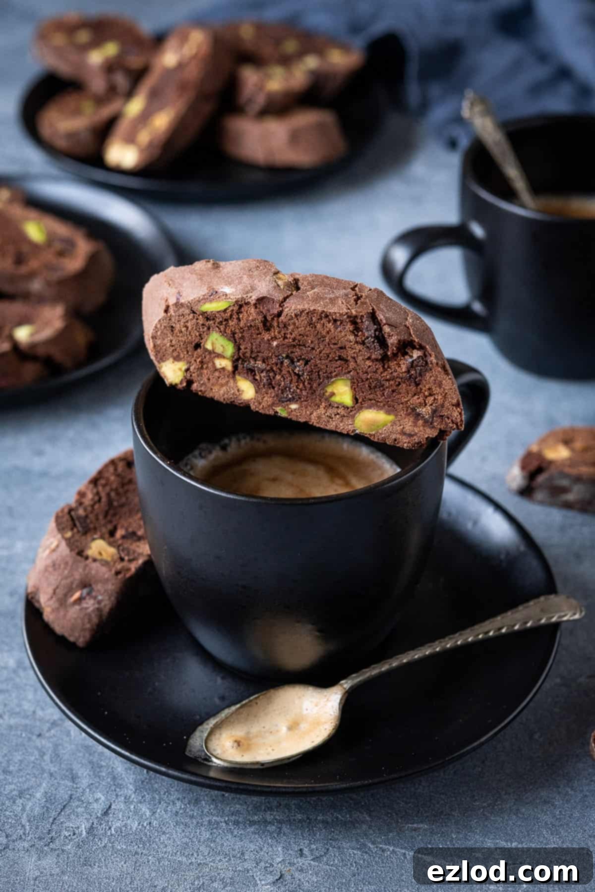A single vegan chocolate pistachio biscotti cookie perfectly balanced on top of a black coffee cup.