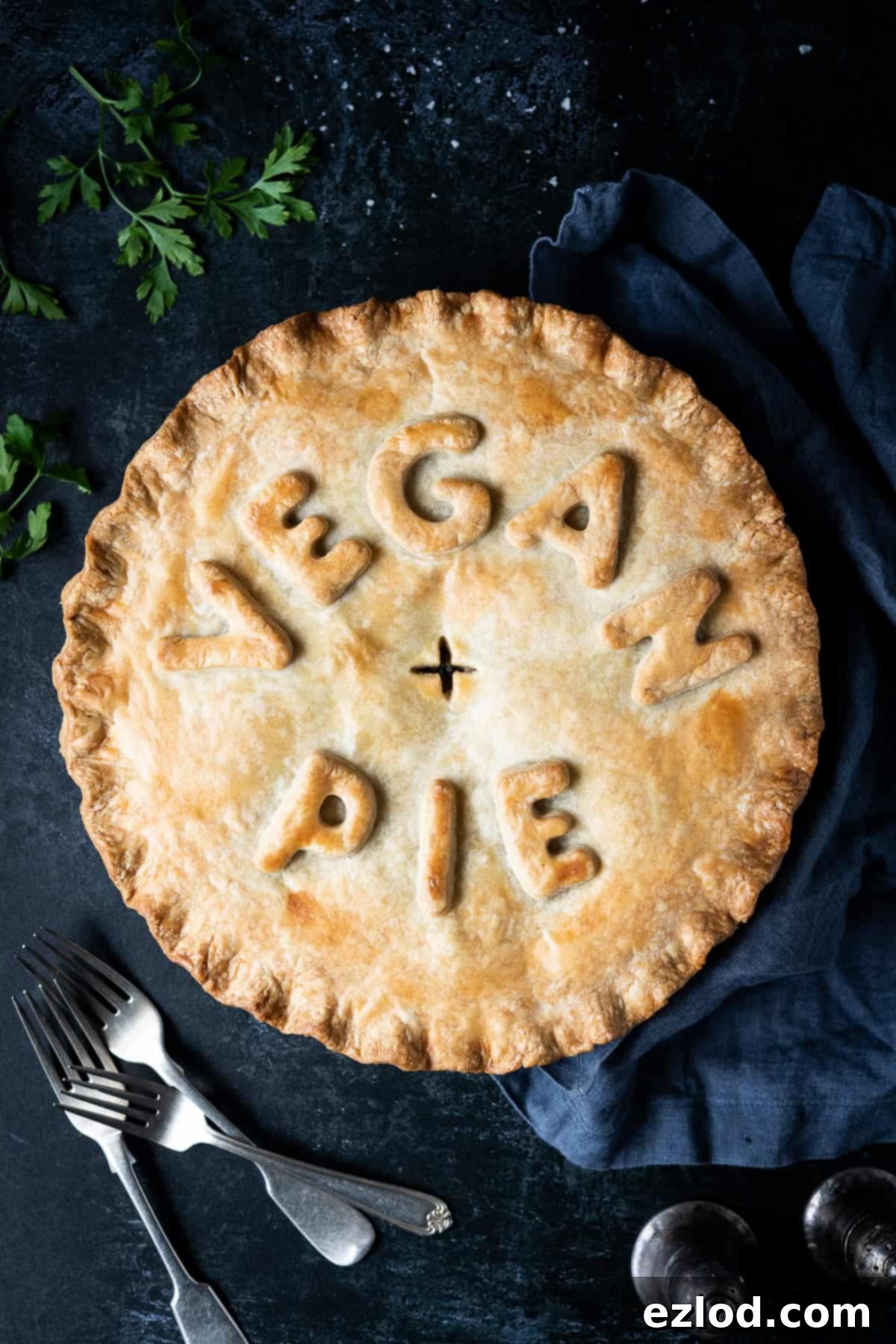 Chickpea, leek and mushroom pie with the words "vegan pie" written on top, on a dark background with forks, parsley, salt and pepper shakers and a grey cloth.