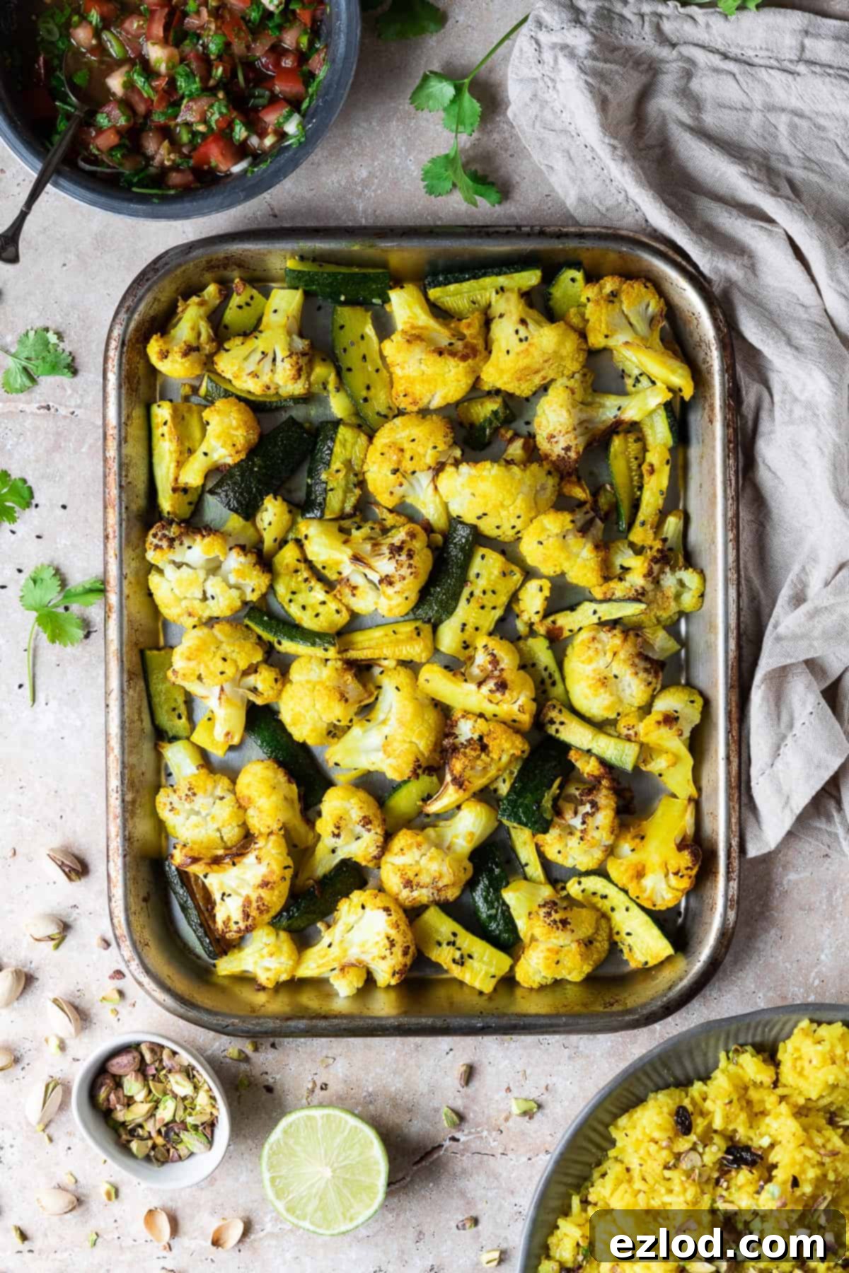A beautifully arranged scene showing a roasting tray of golden-brown spiced cauliflower and courgette, a steaming bowl of bright yellow rice, a vibrant bowl of tomato salsa, a fresh lime half, pistachios, and fresh coriander leaves, all set on a rustic surface.