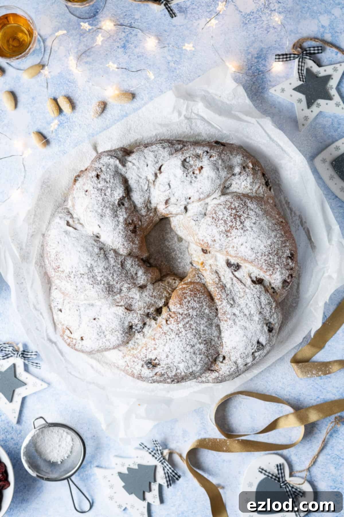 Vegan stollen wreath on a blue background with christmas decorations, christmas lights, gold ribbon, almonds and a glass of rum.