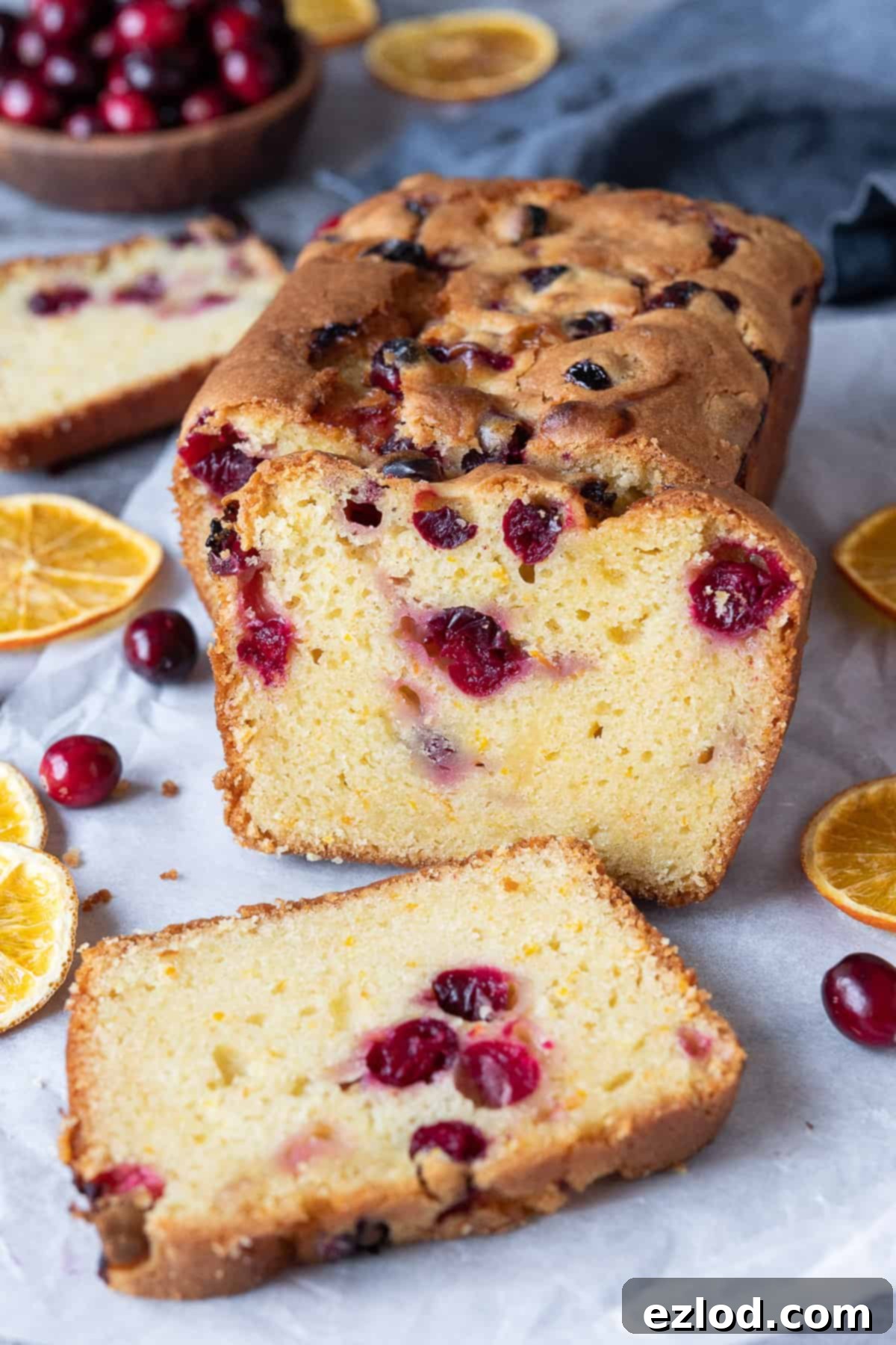 Vegan orange cranberry cake with three slices cut with dried orange slices and a bowl of fresh cranberries in the background.