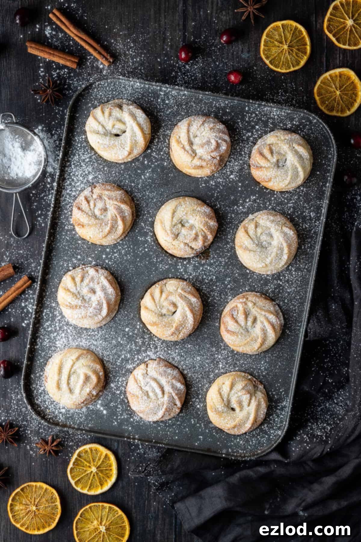 A tray of vegan Viennese whirl mince pies on a dark wooden table, garnished with dried orange slices, cinnamon sticks, cranberries, and star anise. Perfect for a festive holiday spread.