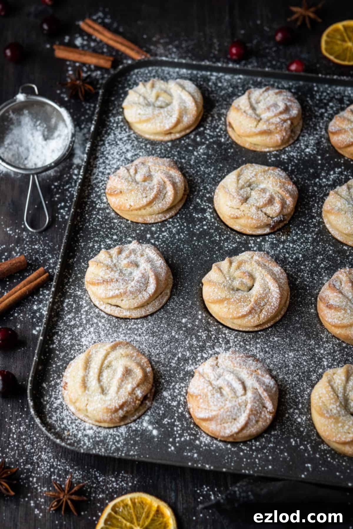 Vegan Viennese whirl mince pies arranged in a metal bun tin on a rustic wooden table, accompanied by a mini sieve with icing sugar, whole spices, and dried orange slices, creating a beautiful holiday scene.