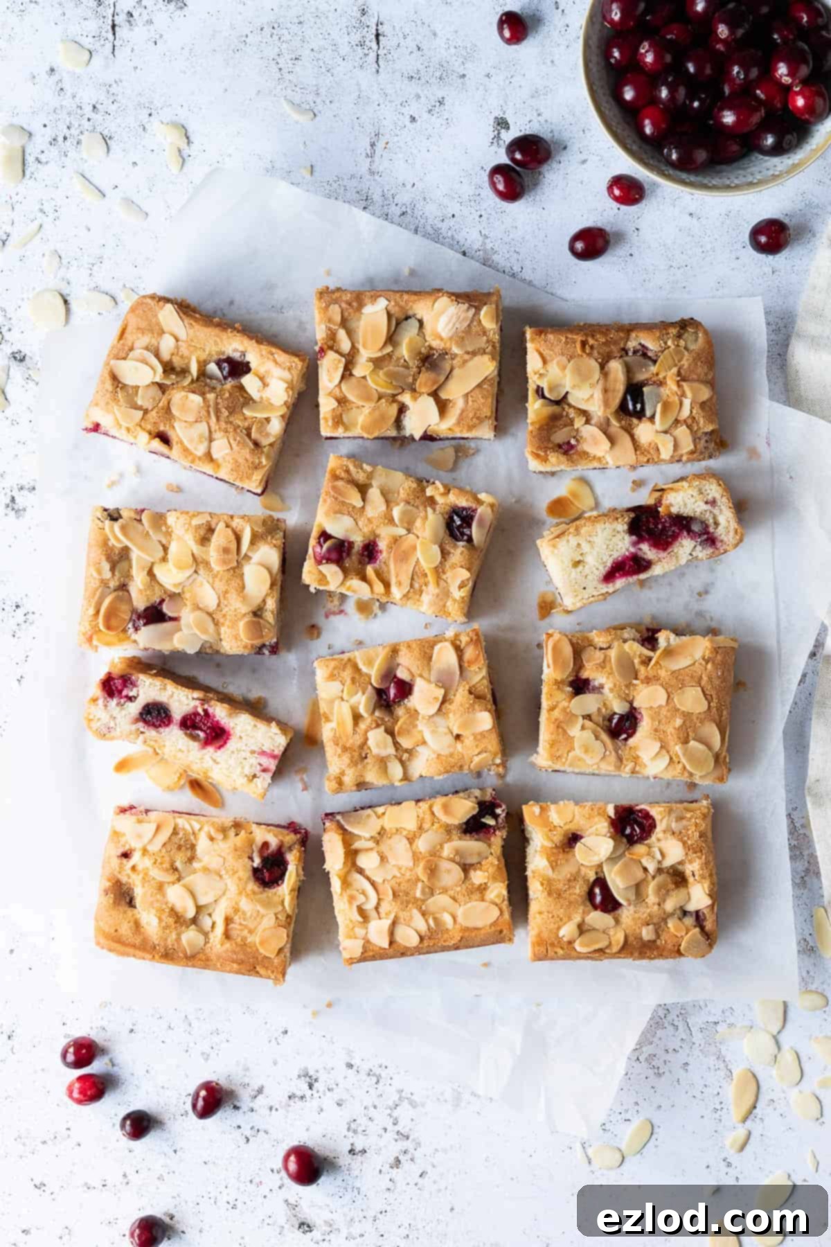 Vegan almond cranberry cake sliced into bars on a sheet of baking parchment on a white background with a bowl of fresh cranberries.