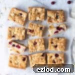Vegan almond cranberry cake sliced into bars on a sheet of baking parchment on a white background with a bowl of fresh cranberries.