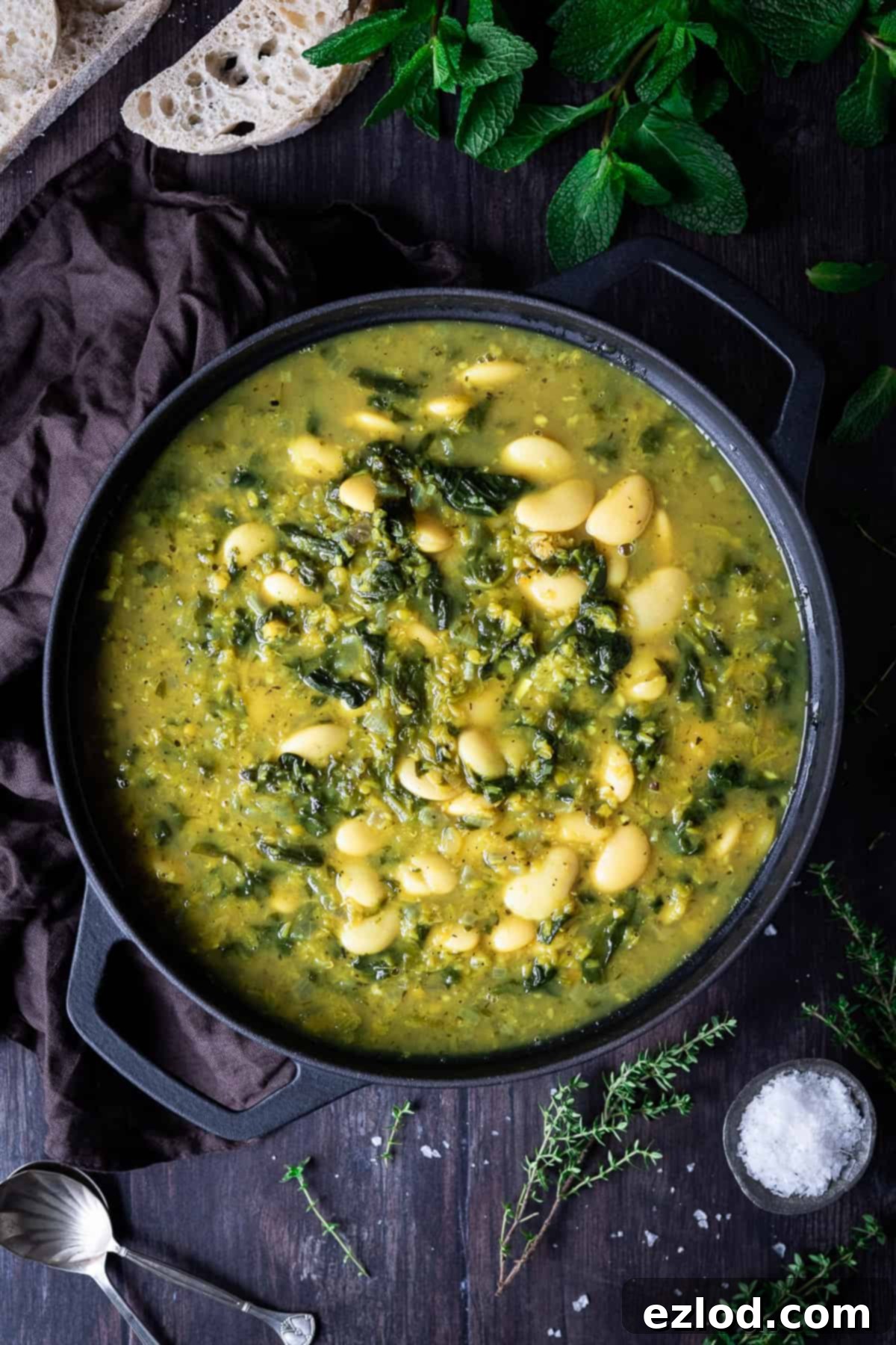 Spinach, lentil and butter bean soup in a black pan on a wooden table with fresh herbs, slices of bread and a bowl of salt.