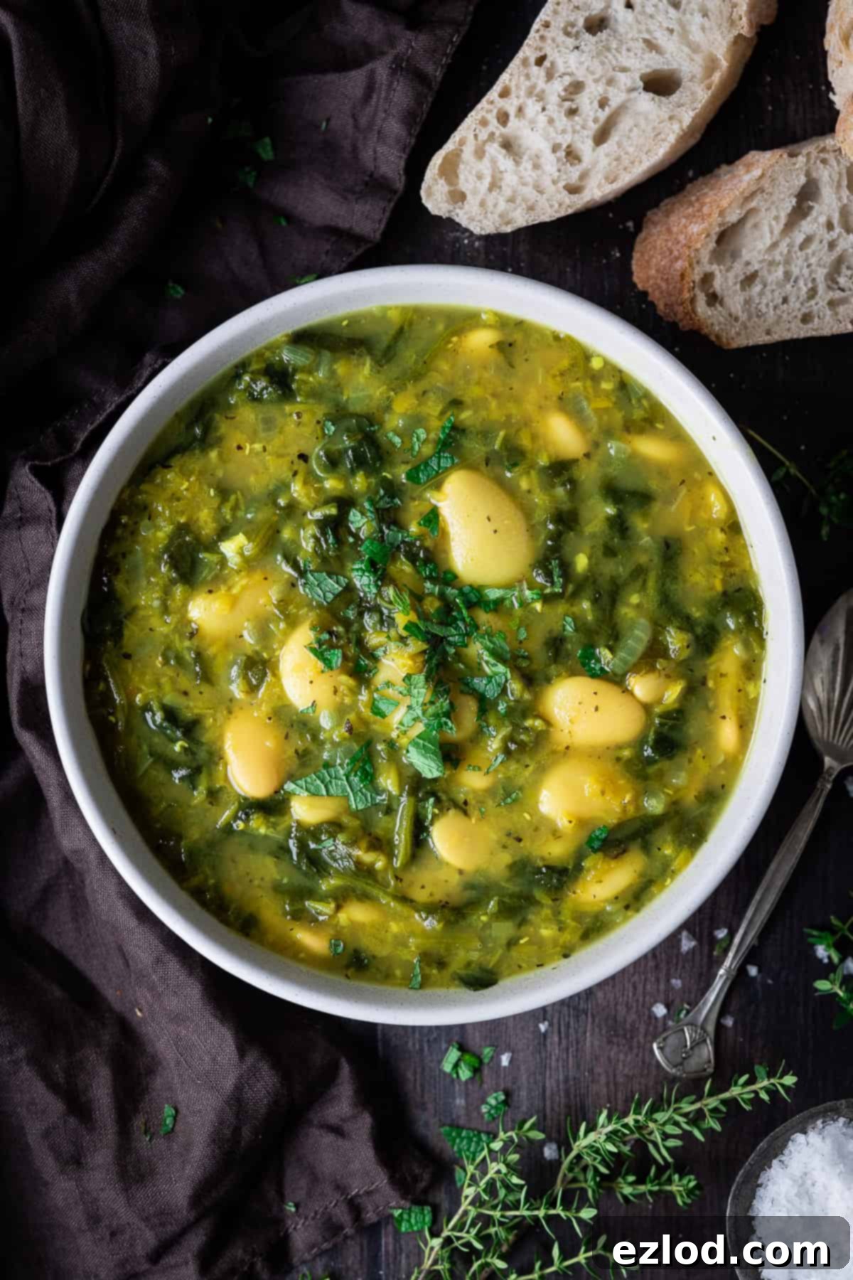 A bowl of soup on a wooden table with a brown cloth, slices of bread and fresh herbs.