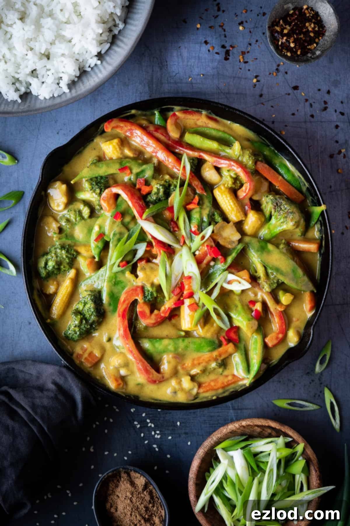 Rich Vegan Chinese curry with a mix of vibrant vegetables in a black skillet, served with fluffy rice, chili flakes, and sliced spring onions on a rustic metal surface, perfect for a cozy plant-based meal.