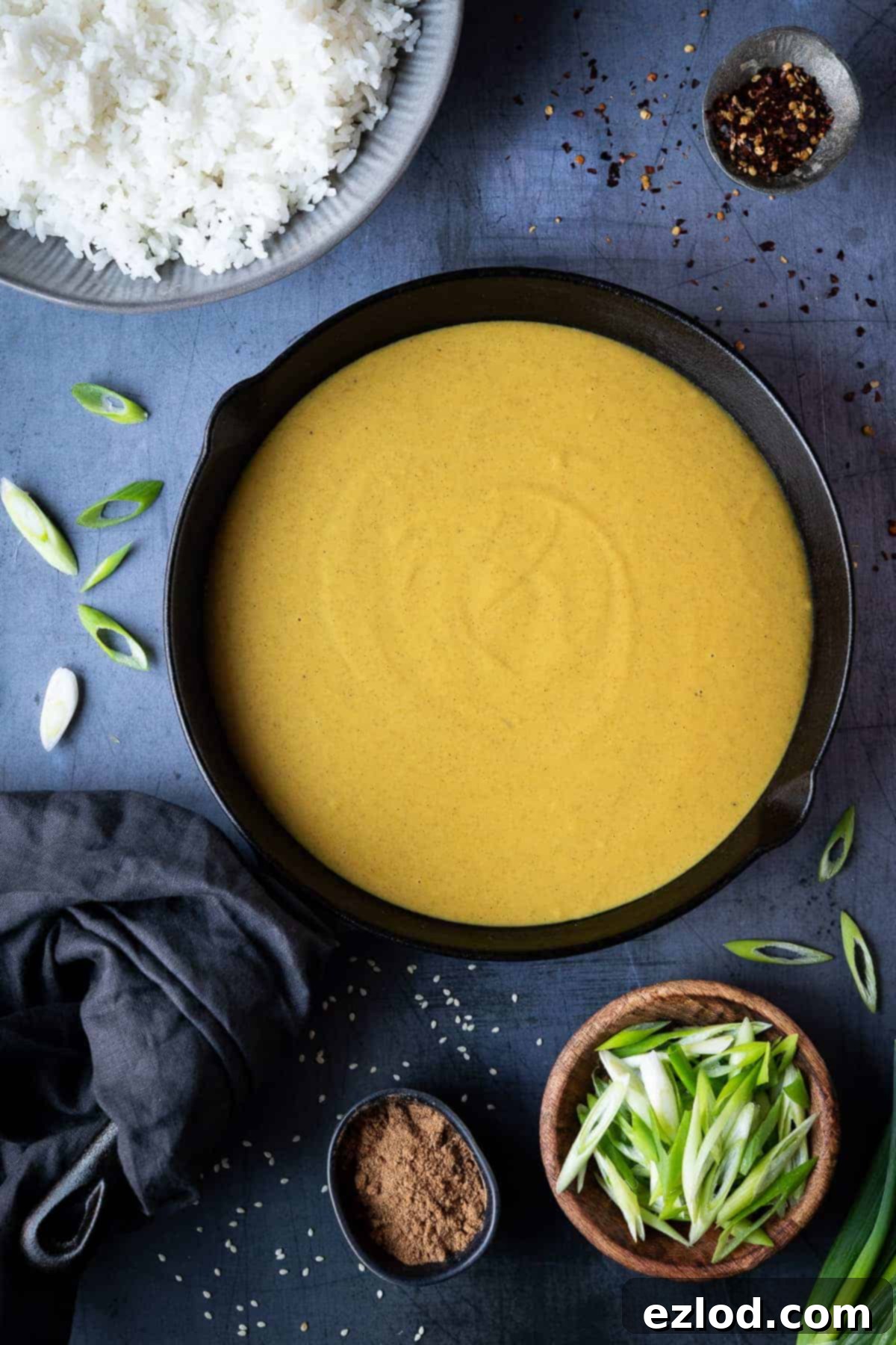 Close-up of creamy vegan Chinese curry sauce simmering in a black skillet on a metal surface, surrounded by a bowl of rice, red chili flakes, and fresh spring onions, highlighting its rich texture and enticing color.
