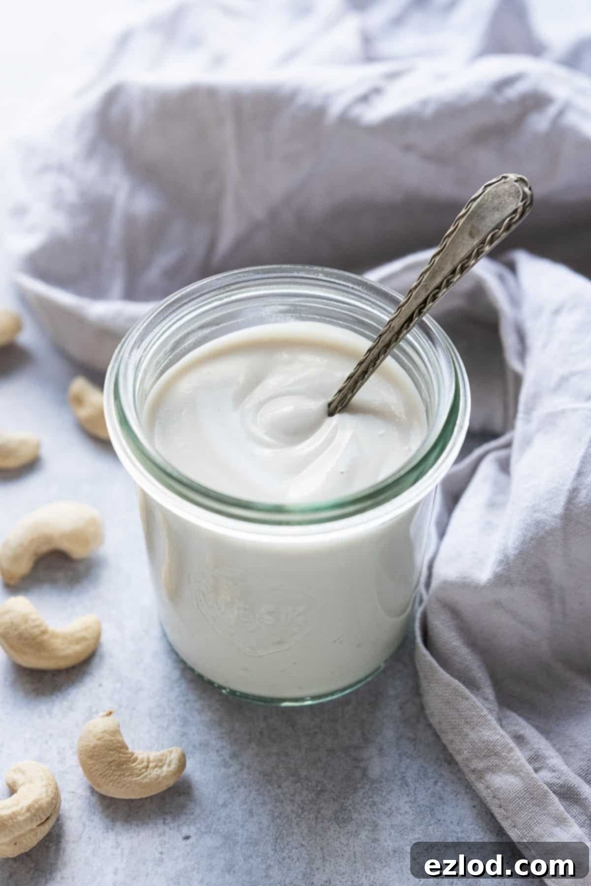 A glass jar of cashew cream with a metal spoon in it with a grey cloth and cashews.