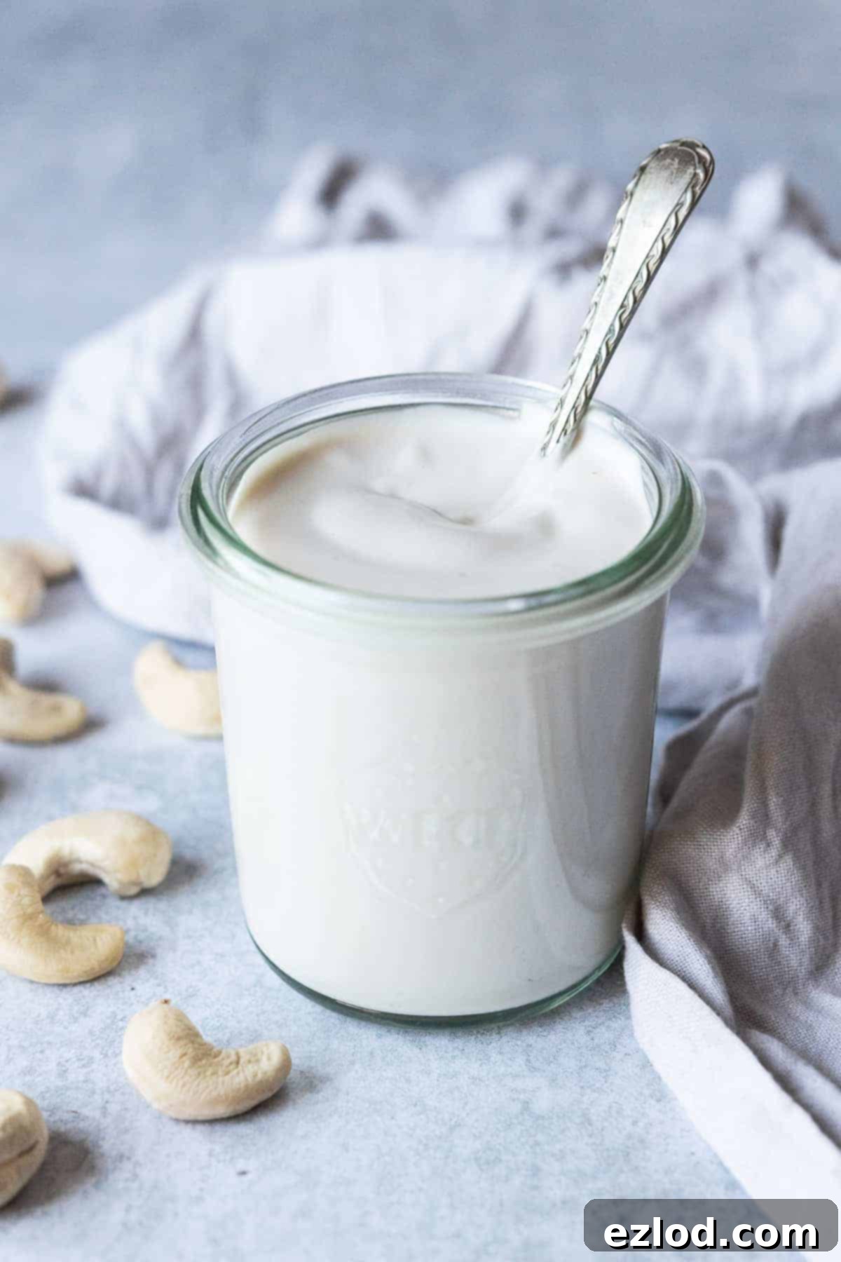 A jar of cashew cream on a grey surface with a scattering of raw cashews and a grey cloth.