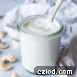 A jar of cashew cream on a grey surface with a scattering of raw cashews and a grey cloth.