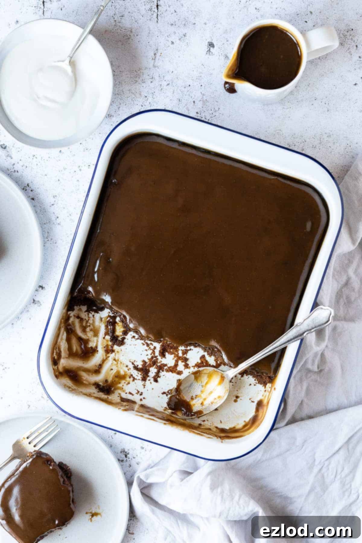 Vegan sticky toffee pudding in an enamel baking dish with several portions scooped out onto plates, with a jug of toffee sauce and a bowl of cream.