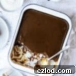 Vegan sticky toffee pudding in an enamel baking dish with several portions scooped out onto plates, with a jug of toffee sauce and a bowl of cream.